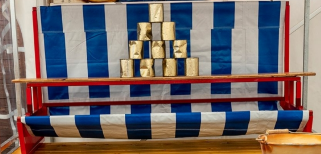 rotes metallgestell mit blau weisser plane. dadrauf ein brett mit10 goldenen verbeulten dosen. so wie vom jahrmarkt.