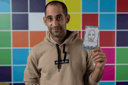 Ein Mann vor einem bunt gekachelten Hintergrund hält in seiner Hand ein vom Draw-Me-Bot erstelltes Porträt in Postkartengrösse hoch.
