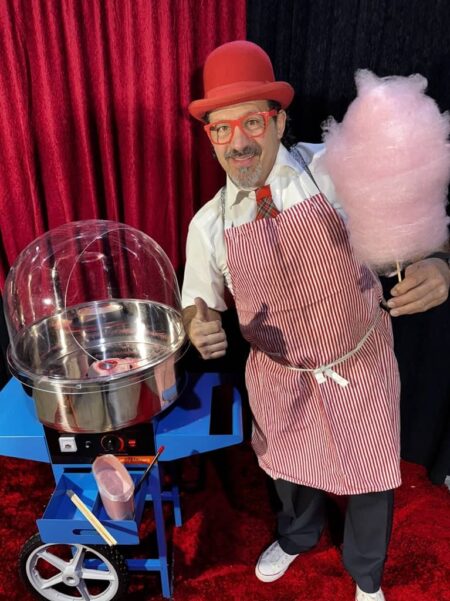clown vor blauer zuckerwattemaschine mit einer rosanen zuckerwatte un ddaume hoch.