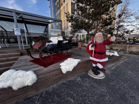 lebensgrosser kunststoff weihnachtsmann vor einem schlitten. er hat einen sack auf der schulter und eine glocke in der hand.