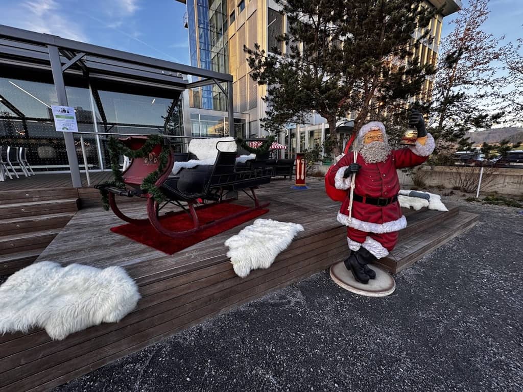 lebensgrosser kunststoff weihnachtsmann vor einem schlitten. er hat einen sack auf der schulter und eine glocke in der hand.
