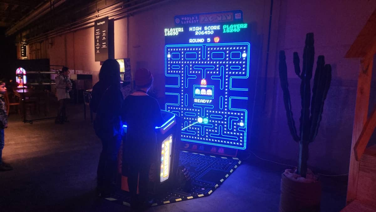 Zwei junge Frauen spielen gemeinsam am XXL Pac-Man Automaten, während ein Junge zuschaut.