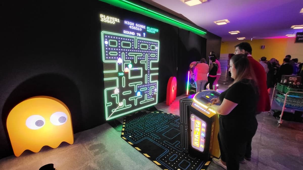 Eine junge Frau und ein junger Mann spielen gemeinsam an der XXL-Pac-Man-Konsole. Neben dem großen LED-Display, befinden sich zwei aus dem Spiel bekannten gelbe und eine rote Gespensterfiguren in Übergröße.